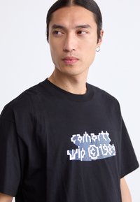 Mand med langt mørkt hår, der er sat tilbage, iført en sort Carhartt t-shirt med hvid grafisk tekst, som ser til venstre mod en ensfarvet lys baggrund.