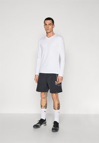 Camisa blanca de manga larga con cuello en V, combinada con pantalones cortos negros que presentan detalles en amarillo y gris, usados con zapatos negros y calcetines blancos.