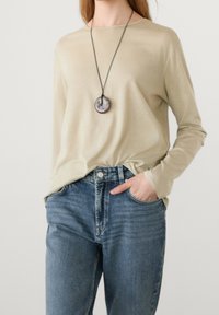 Pitkähiha beige paita, joka on valmistettu pehmeästä kankaasta, yhdistetty korkeavyötäröisten sinisten farkkujen kanssa. Pyöreä riipuskaulaketju lisää yksityiskohtia.