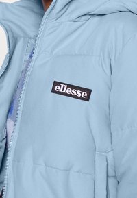 Jasnoniebieska kurtka puchowa z kapturem, z zamkiem błyskawicznym i haftowanym czarno-białym logo "ellesse" na klatce piersiowej.
