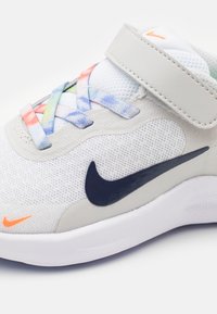 Zapatilla Nike con una parte superior de malla blanca, logotipo swoosh en navy, cordones coloridos (azul, rojo, verde) y una correa de Velcro gris. Suela de goma.