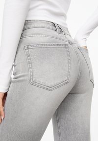 Jean en denim gris à coupe ajustée, avec deux poches arrière et une texture légèrement délavée. Associé à un haut blanc à manches longues.