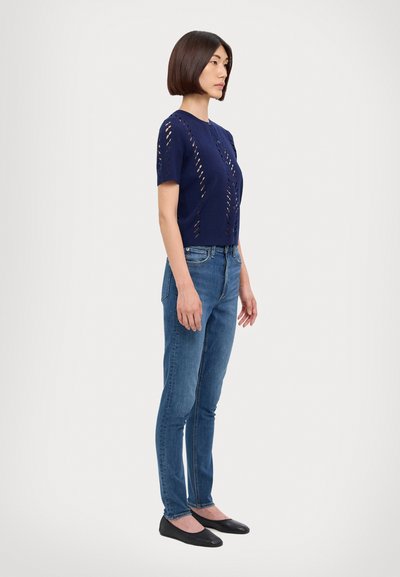 Top corto a maglia blu navy con motivi a taglio, abbinato a jeans aderenti azzurri e ballerine nere. Vista laterale, modello in piedi.