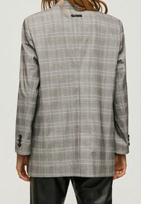 Blazer de cuadros gris con un corte relajado, que presenta detalles en negro, puños de un solo botón y una textura suave. La vista trasera muestra claramente el patrón de cuadros.