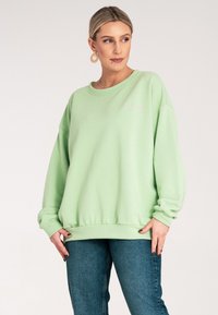 Jasnozielony, oversize'owy sweatshirt z okrągłym dekoltem, ściągaczami na mankietach oraz małym, różowym haftowanym napisem na klatce piersiowej.