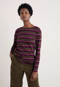 Dugi rukav prugasti top s horizontalnim prugama u ružičastoj, zelenoj i crnoj boji, s brodom izrezom, uparen s teksturiranim maslinasto zelenim baršunastim hlačama.