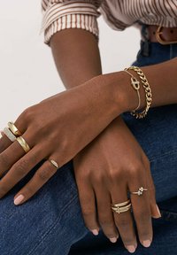 Guld ringe i forskellige design, herunder teksturerede og glatte overflader, lagdelt på hænder med et guld kædearmbånd mod denim.
