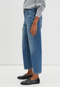 Personne portant un jean cropped bleu délavé, un haut à manches longues gris clair et des mocassins en cuir noir, se tenant de côté devant un fond blanc.