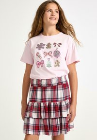 Lyserød t-shirt med farverige, festlige grafiske designs. Rød og hvidternet nederdel med flæser og elastisk talje.