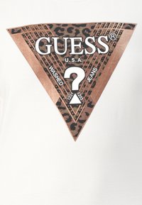 Hvid t-shirt med et brunt leopardprint trekantet logo med teksten "GUESS U.S.A." og "WASHED JEANS", dekoreret med gyldne nitter.