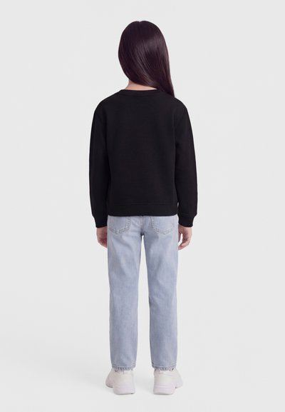 Barn som står med ryggen mot betrakteren, iført svart genser, lyseblå jeans og hvite joggesko mot en ensfarget bakgrunn.