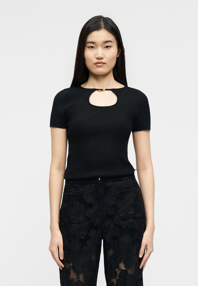 Γυναίκα με μακριά σκούρα μαλλιά φοράει μαύρο ribbed κοντομάνικο τοπ με άνοιγμα στο μπροστινό μέρος και ψηλόμεσα μαύρα λινά δαντελένια παντελόνια με λουλουδάτο μοτίβο, στέκεται μπροστά σε άσπρο φόντο.