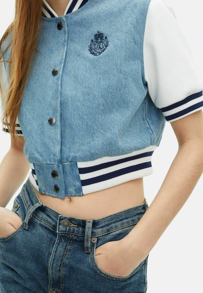 Skórzana kurtka z denimu z niebieskiego materiału, białymi rękawami i granatowym, prążkowanym kołnierzem. Posiada zapięcie na guziki i haftowane logo.