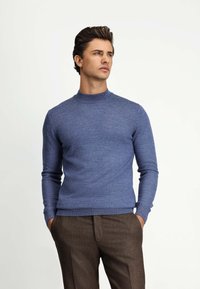 Niebieski sweter robiony na drutach z wysokim kołnierzem, długimi rękawami i prążkowanymi wykończeniami, w połączeniu z brązowymi spodniami typu chinos. Gładka tekstura, minimalistyczny design.
