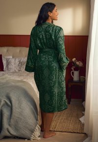Una donna in una vestaglia verde a fantasia è in piedi scalza accanto a un letto con una coperta grigia, un tavolino di legno tiene dei fiori e una tazza in una camera da letto accogliente.