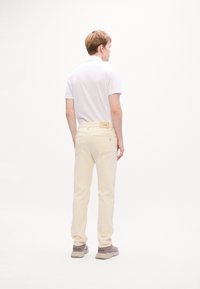 Homme portant un polo blanc à manches courtes, un jean couleur crème et des baskets beiges, se tenant de dos contre un fond blanc uni.