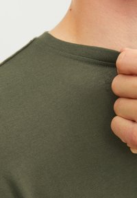 Olijfgroen katoenen T-shirt met een ronde hals, licht ribbelige textuur, hand die de kraag vasthoudt, zijnaad zichtbaar.