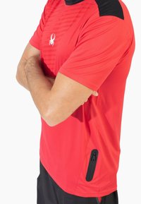 Rood sportshirt met zwarte accenten, korte mouwen en een ritssluiting aan de zijkant. Heeft gestructureerde strepen aan de voorkant en een logo.