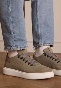 Green suede sneakers with grey laces and metal eyelets, featuring a white rubber sole. Worn with frayed light blue jeans and white socks.