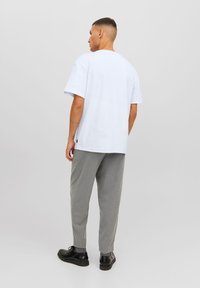 Camiseta de algodón blanca de manga corta, corte relajado, y una pequeña etiqueta de la marca cerca del dobladillo, combinada con pantalones grises con patrón y zapatos negros.