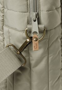 Lichtgroene gewatteerde tas van dichtbij, met een YKK-rits, messing hardware en een leren accentlabel met een debossed "J." Geweven band zichtbaar.