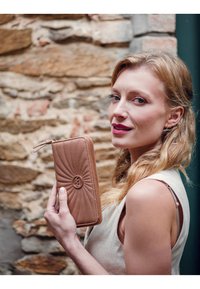 Brown quilted wallet with a gold logo, featuring a zip closure and textured design. The fabric is smooth with a subtle sheen.