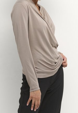 Maglietta beige a maniche lunghe con scollatura drappeggiata e vestibilità rilassata. Realizzata in tessuto morbido, abbinata a pantaloni neri su misura. Texture liscia, senza motivi visibili.