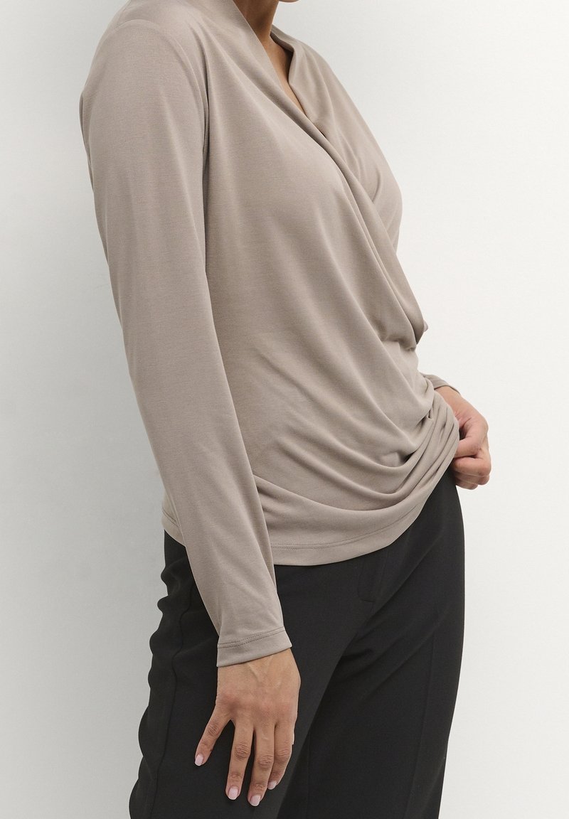 Beige Langarmshirt mit drapierter Halslinie und lockerer Passform. Hergestellt aus weichem Stoff, kombiniert mit schwarzen, maßgeschneiderten Hosen. Glatte Textur, keine sichtbaren Muster.