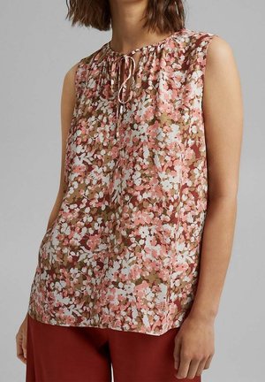 Blouse sans manches à fleurs roses, blanches et marron, avec un col rond et un détail à nouer, portée avec un pantalon couleur rouille.