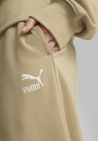 Bežs sporta krekls ar ribotām piedurknēm, ar sānu kabatu un izšūtu balto Puma logotipu priekšpusē, gluda tekstūra ar minimālistisku dizainu.
