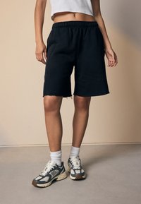 Schwarze Baumwollshorts mit lockerem Sitz und ausgefranstem Saum, kombiniert mit grauen und weißen Sneakers sowie weiß gerippten Socken.