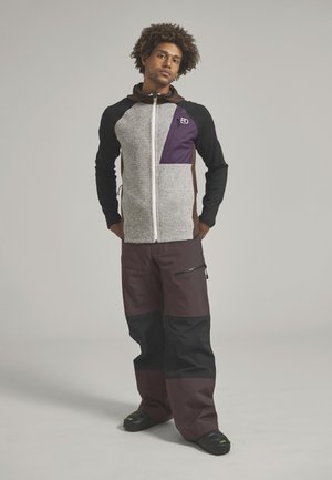 Mann mit lockigem Haar, der einen mehrfarbigen Kapuzenpullover mit Reißverschluss und braune Hosen mit schwarzen Knieschonern trägt, steht mit den Händen hinter dem Rücken.