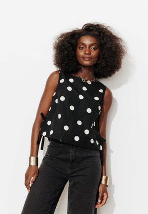 Femme aux cheveux volumineux et bouclés portant un haut noir sans manches à gros pois blancs, un pantalon noir taille haute, des bracelets dorés et un collier.