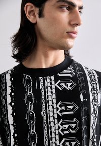 Čierne grafické tričko s odvážnymi bielymi vzormi retiazok a textu. Rukáv s klasickým golierom a krátkymi rukávmi, vyrobené z hladkej textúrovanej tkaniny.