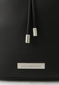 Juoda odinė rankinė su dviem juodais raišteliais, baigiančiais sidabriniais dangteliais, ir sidabrine plokštele, ant kurios įspaustas prekės ženklas „KARL LAGERFELD“.