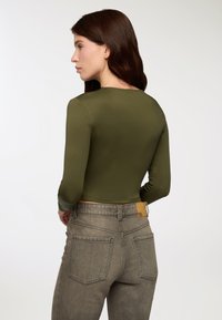 Oliwkowa bluzka z długim rękawem o dopasowanym kroju, zestawiona z szarymi dżinsami z skórzaną naszywką na pasku i tylnych kieszeniach.