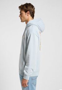 Felpa azzurra chiara con tasca frontale, cappuccio con cordino e grafica del logo. Polsini e orlo a coste per una vestibilità aderente. Materiale in cotone morbido.