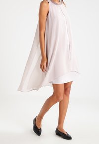Robe sans manches rose pâle avec un design de cape fluide, décolleté orné et doublure intérieure blanche contrastante ; portée avec des ballerines noires.