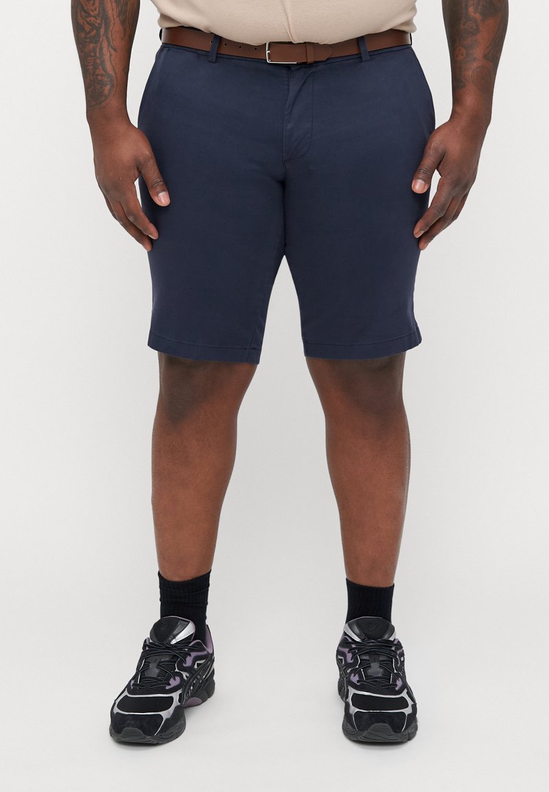 Marineblå shorts med lige skæring og ombukket kant, med en flad front og sidelommer, parret med sorte og lilla sneakers.