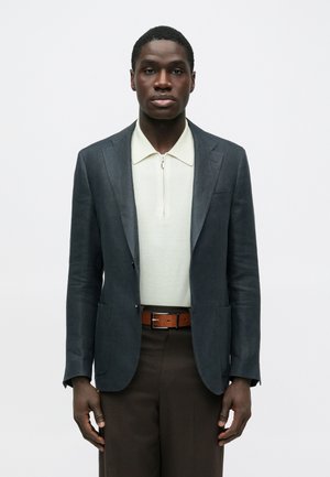 Άνδρας φορώντας σκούρο γκρι σακάκι πάνω από μπλούζα πόλο με φερμουάρ σε κρεμ απόχρωση, καφέ παντελόνι και καφέ δερμάτινη ζώνη, στέκεται μπροστά σε απλό λευκό φόντο.