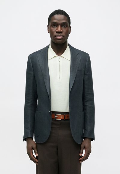 Homme portant un blazer gris foncé sur un polo à fermeture éclair crème, un pantalon marron et une ceinture en cuir marron, debout devant un fond blanc uni.