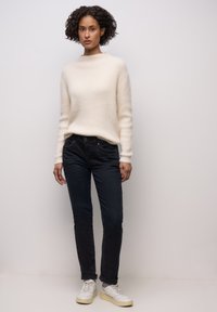 Kremowy, fakturowany sweter z wysokim dekoltem, zestawiony z ciemnymi dżinsami i białymi trampkami, z subtelnymi detalami wzorniczymi i przeszyciami.