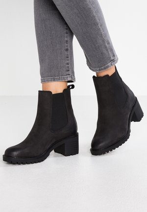 Person iført sorte læderankelstøvler med blokhæl og elastiske sider, kombineret med grå knækkede jeans mod en hvid baggrund.