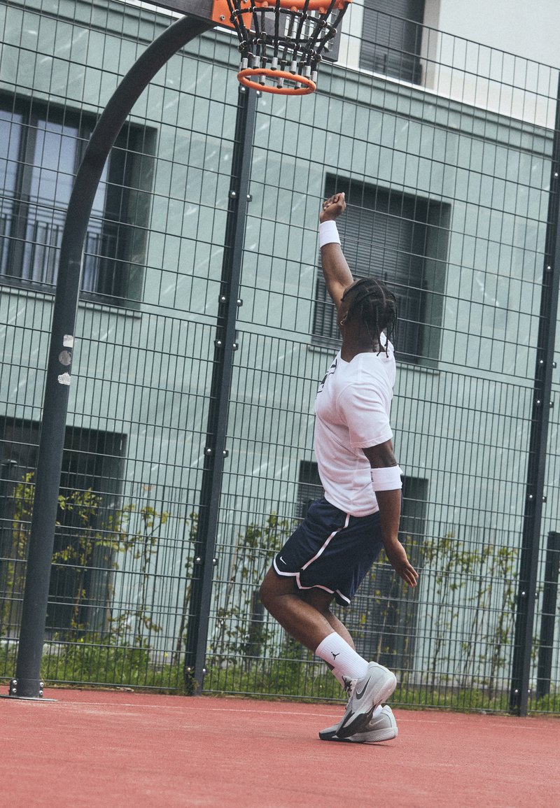 Un athlète portant un T-shirt blanc et un short bleu marine saute pour tirer au basket. Il porte des baskets noir et blanc ainsi que des bracelets de poignet, sur un terrain rouge.