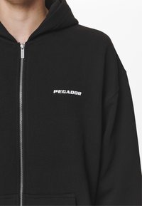 Felpa nera con zip realizzata in tessuto morbido, dotata di tasca a marsupio, un ampio cappuccio e la scritta "PEGADOR" in bianco sul petto.