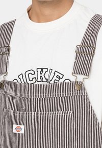 Salopette rayée marron et blanche avec des clips en métal ; portée par-dessus un t-shirt blanc avec un logo imprimé en noir. Détail notable : une poche avant.