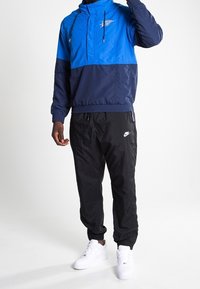 Niebieski i granatowy kurtka typu windbreaker z wysokim kołnierzem i sznurkiem, w połączeniu z czarnymi zwężanymi spodniami dresowymi i białymi sneakersami. Widoczne logo Nike.