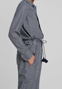 Randig byxdress med krage, långa ärmar, midjeband med tofsar och sidofickor. Tillverkad av ett lätt tyg i marinblått och vitt.