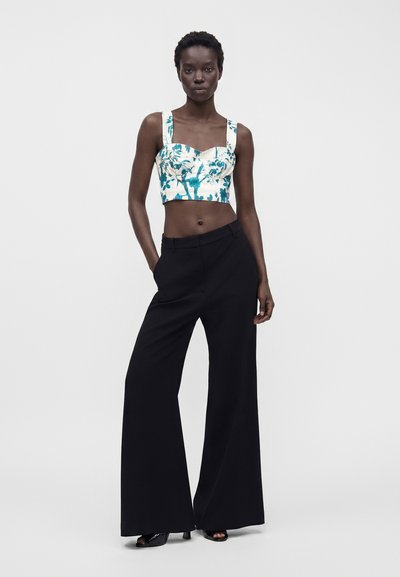 Femme portant un top court fleuri blanc et bleu sarcelle avec des larges bretelles, un pantalon noir à jambes larges et des talons noirs à bouts ouverts, debout, une main dans la poche.