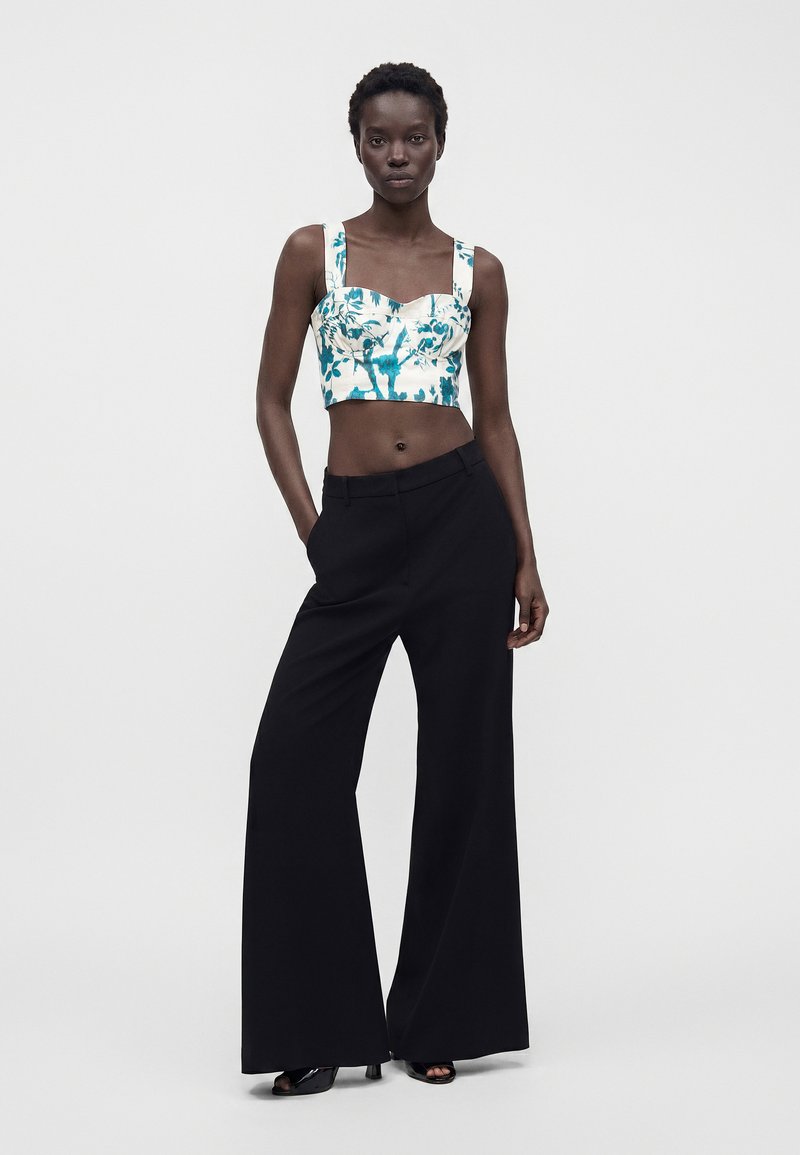 Woman wearing a white and teal floral cropped top with wide straps, black wide-leg pants, and black open-toe heels, standing with one hand in pocket.